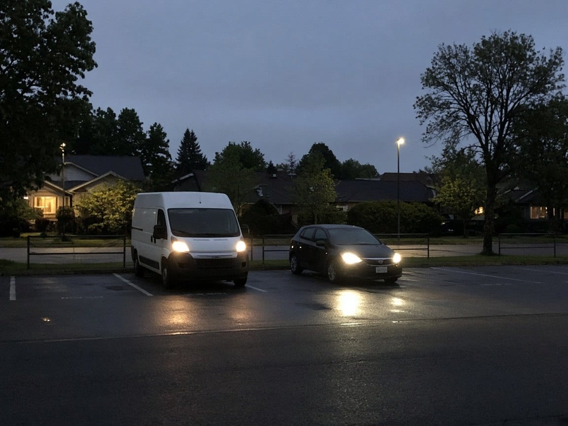A van and a car with headlights on in a lot at dusk
